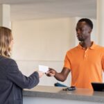 A customer hands a card across a dealership service counter to an advisor in an orange polo, car keys resting beside them