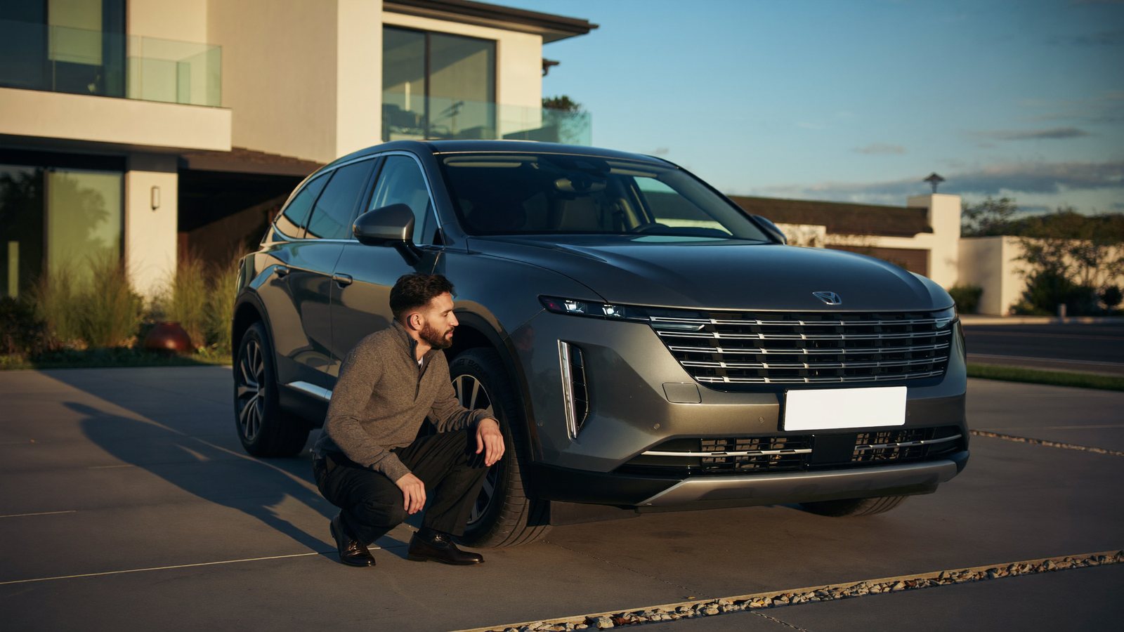 A man crouches beside the front wheel of his late-model crossover in his driveway at dusk, checking the tire