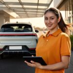 A service advisor in an orange polo stands in a dealership service drive as a customer's crossover pulls away down the lane