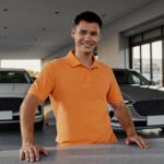 A smiling service advisor in an orange polo stands behind the write-up counter in a dealership service drive, with customer vehicles waiting in the lane behind him
